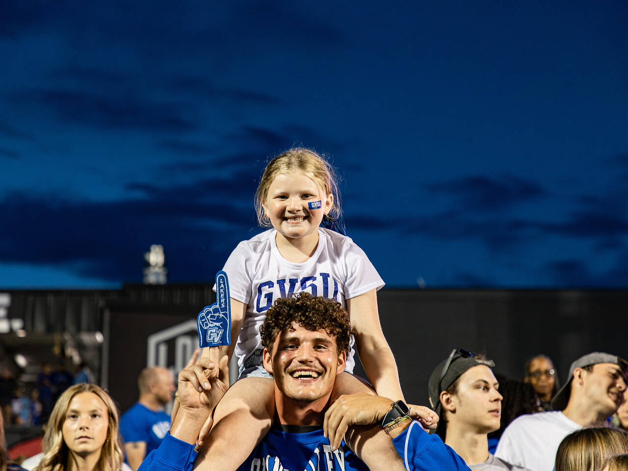 A young fan sits on the shoulders of a supporter at a sports event, both smiling under a darkening sky.